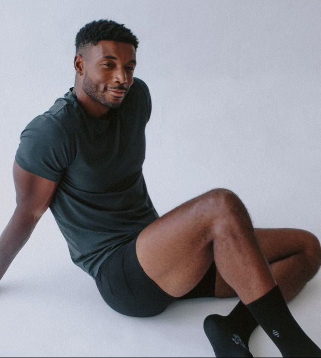 Man sitting on a white floor wearing a dark green t-shirt and black shorts against a light gray background